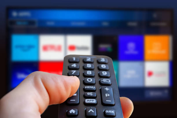 Hand holding a remote control in front of a TV screen displaying various streaming service apps.